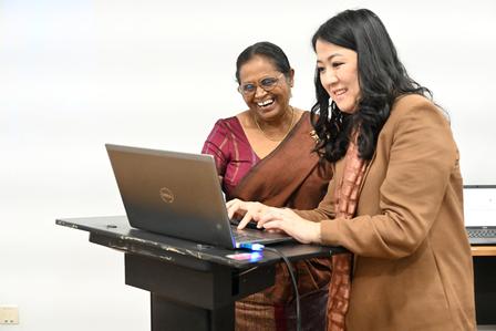 Two women stand at a podium with a laptop, giving a presentation.
