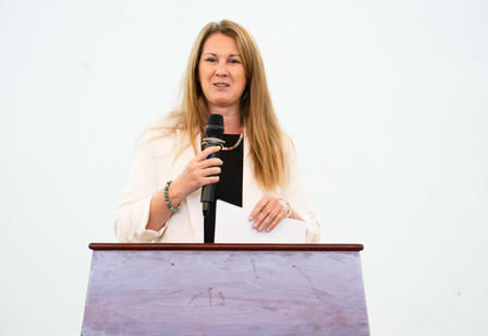 Person behind a purple lectern, microphone in hand, white background.