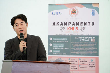 Man at podium delivering a speech, with a banner displaying event details behind him.