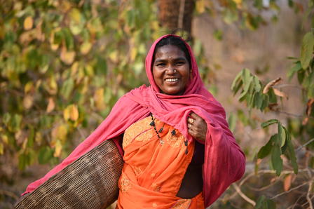Person in orange dress with pink scarf holding a woven basket outdoors among trees.