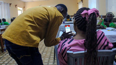 Man leaning toward a girl with braids in pink beside a pink wheelchair in a busy hall.
