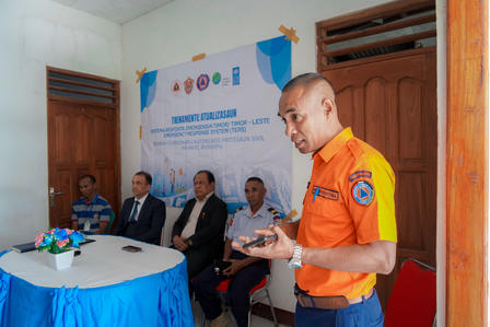 Man in orange uniform presenting to a panel at a formal meeting with a blue banner in the background.