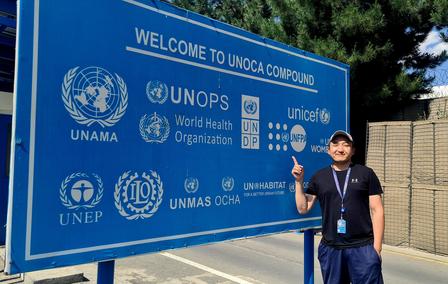 Photo: man with a lanyard stands in front of a large blue UN banner outdoors.