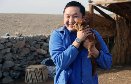 Man in a blue jacket holding a small brown animal near his shoulder outside a wooden shelter.