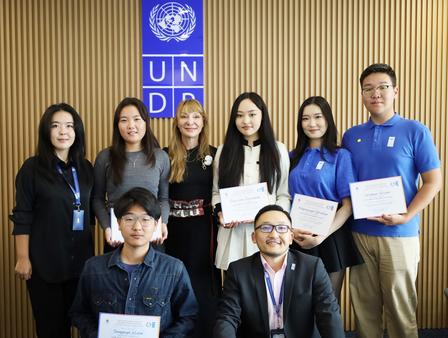 Group photo of students and a man posing with certificates in front of a UN banner.