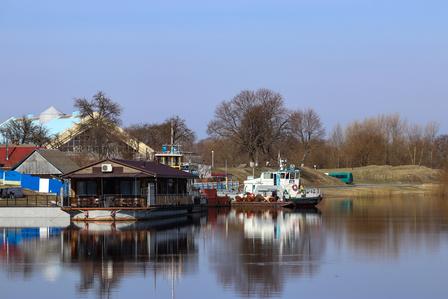 Calm lake with moored boats and wooden buildings along the shore, reflected in the water.