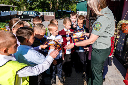 Photograph: Group of children gathered around a uniformed woman handing out items in a sunny outdoor setting.