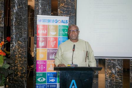 A man stands at a podium with a microphone, presenting in front of colorful SDG banners.
