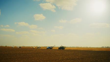 Two tractors plowing a wide brown field under a bright blue sky with the sun.