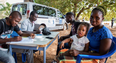 Group of people, including children, drawing at a table outdoors; a white van and trees in the background.