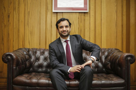 Man in a dark suit and red tie sits on a brown leather sofa in a wood-paneled office.
