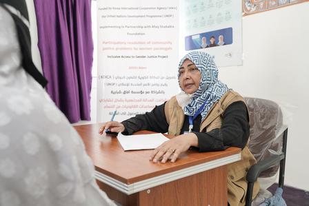 Photograph of a student writing at a desk in a classroom, wearing a patterned headscarf.