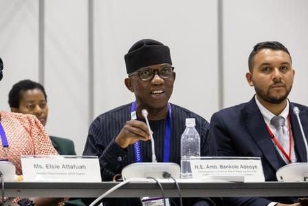 Three panelists seated at a conference table with nameplates, microphones, and water.
