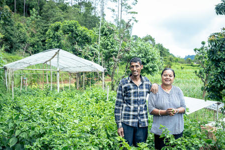 Two smiling people stand amidst lush greenery, with a cultivation structure in the background.