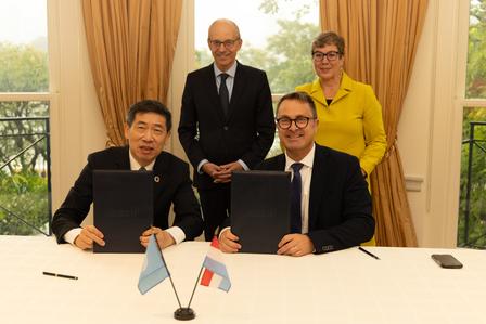 Five people in a formal signing ceremony; two men sign documents at a table with small flags, others stand behind.