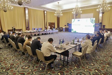 Large conference room with participants seated around U-shaped tables, projection screen at front.
