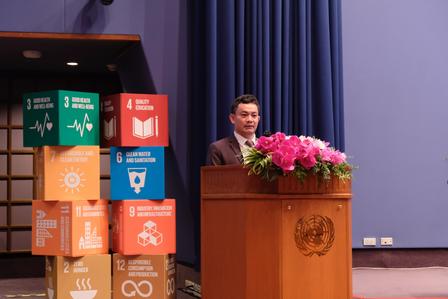 Photograph of a man speaking at a podium with a flower arrangement; SDG blocks on the side.