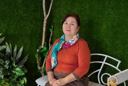 Woman sits in a garden on a white circular chair, wearing an orange top and a colorful scarf.