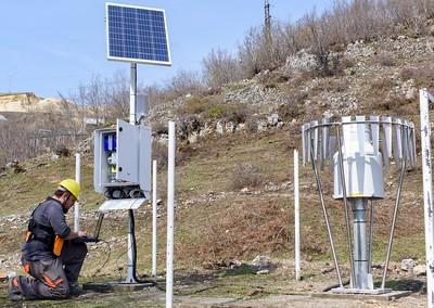 Field technician kneels beside a solar panel and weather sensors on a dry, rocky hillside.