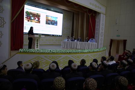 Auditorium stage with a panel at a long table; speaker at podium, audience wearing caps.
