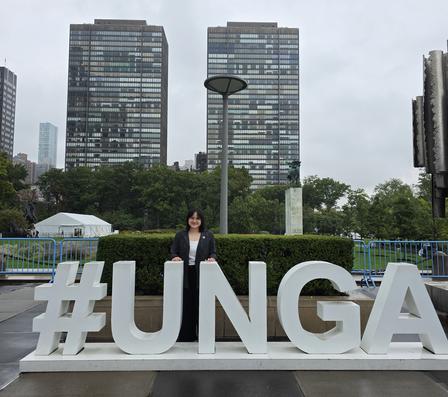 Woman stands behind large white #UNGA letters on a plaza with hedges and tall buildings nearby.