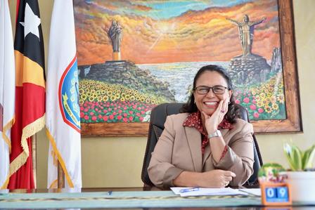 Government official at desk in an office with flags, a plant, and a colorful painting.