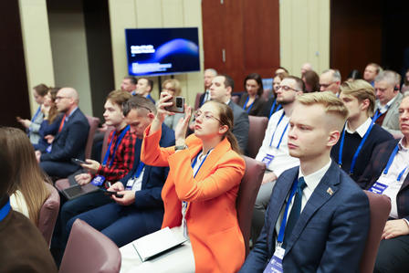 Conference audience; woman in orange blazer speaks into handheld microphone.