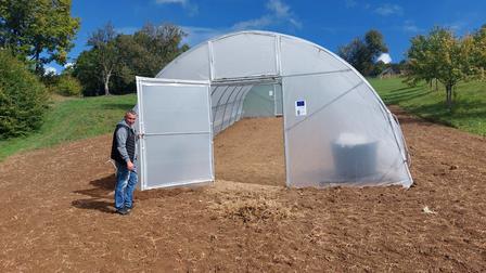 Person stands beside a white hoop-house greenhouse in a dirt field under a blue sky.
