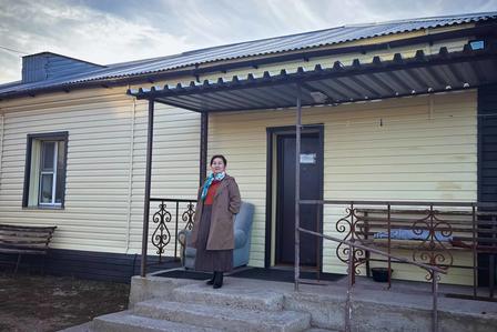 Person in a long brown coat stands on steps before a pale-yellow building with a bench.