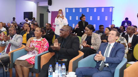 Diverse audience seated in a conference hall, watching a presentation on stage with blue backdrop.