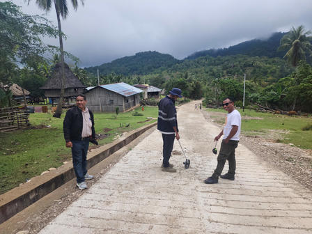 Three men stand on a rural road in a hillside village with lush green hills.