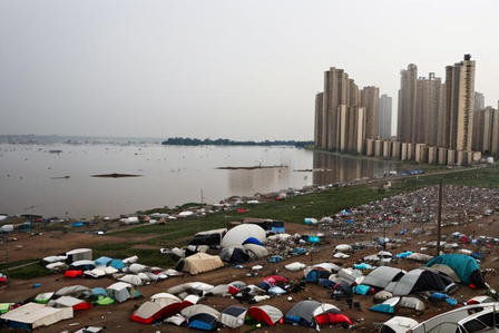 Crowded seaside campsite with colorful tents; calm water and distant city skyline.