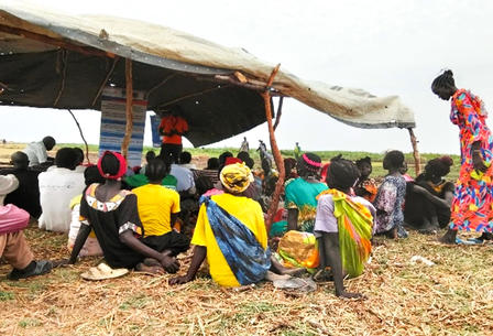 People in colorful attire sit on the ground under a tarp, listening to a speaker.