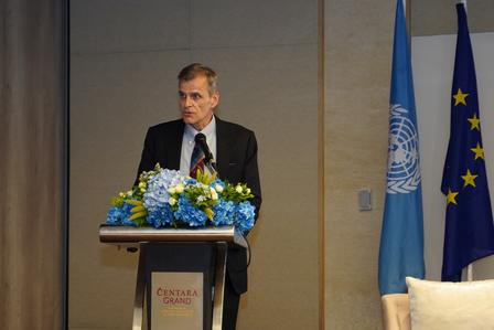 Photograph of a man speaking at a podium with blue flowers, UN and EU flags in the background.