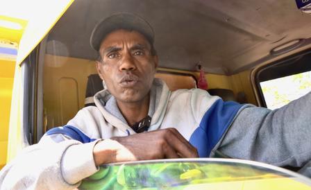 Person in a gray hoodie sits in a yellow bus cabin, hands folded over a green patterned surface.