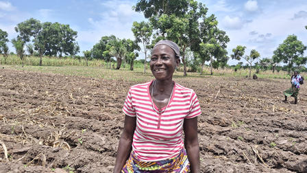 Person in pink striped shirt and colorful belt stands in a plowed field with trees and blue sky.
