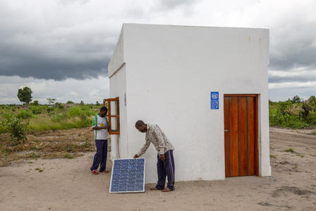 Two men stand by a small white building with a wooden door in a dusty field; a solar panel by the wall.