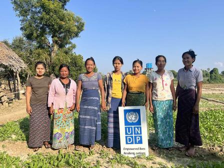 Group photo of women in traditional dress standing in a rural village beside a UNDP sign.