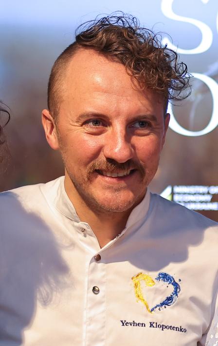 Person in white polo with blue crest logo on chest, standing before a blue backdrop.