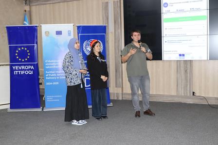 Three people on a conference stage, blue EU banner to the left and a projection screen behind.