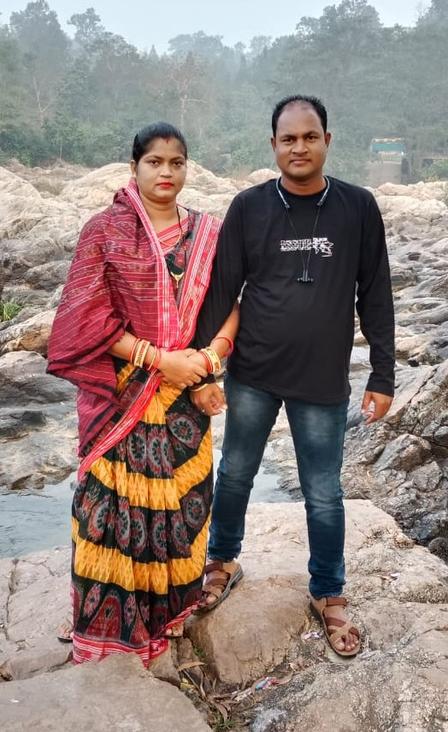 Two people stand on a rocky riverbank; woman in colorful traditional dress, man in black shirt.