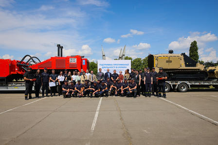Group of people posing in front of a red steam locomotive, tan armored vehicle, and banner outdoors