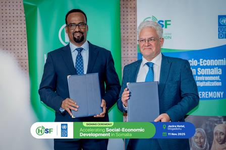 Two men in suits hold documents on stage at a ceremony, with green flag and blue banners behind.