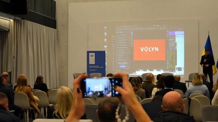 Conference audience watches a screen displaying VOLYN; foreground person snaps a photo.