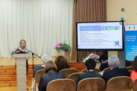 Speaker at podium presenting to audience in a conference hall with a slide on screen.