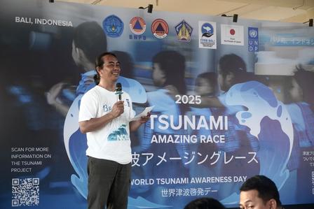 Photo of a man in a white T-shirt speaking on stage in front of a blue tsunami awareness banner.