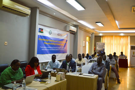 Conference room with attendees seated at long tables; banner on the wall behind.
