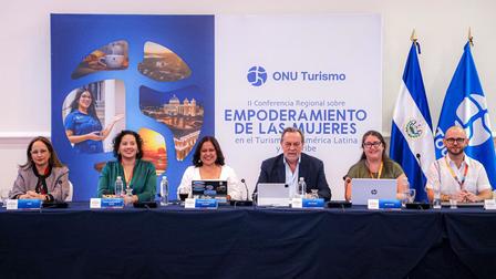 Photograph of six people seated at a conference panel on a stage, with a blue banner and flags behind them.
