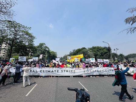 Large crowd of protesters marching on a sunny street with signs and a banner; camera in foreground.