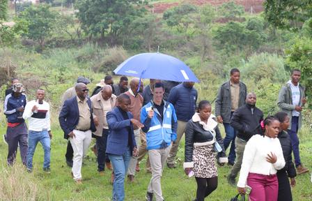 Group of people standing in a grassy field with trees in the background; one person holds a blue umbrella.